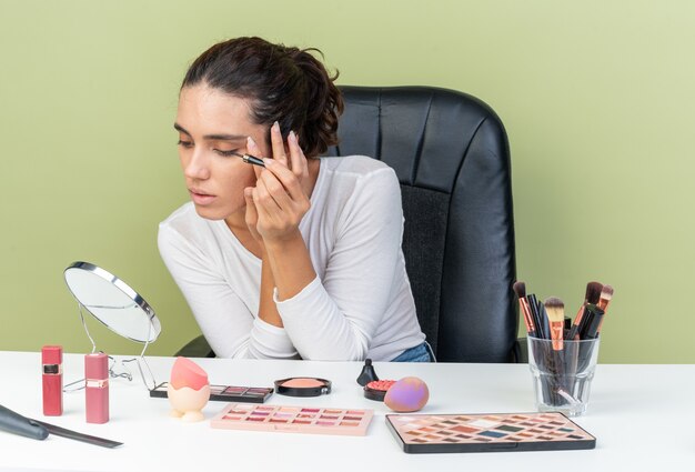 Foto grátis mulher bonita caucasiana confiante sentada à mesa com ferramentas de maquiagem, aplicando delineador, olhando para o espelho isolado na parede verde oliva com espaço de cópia