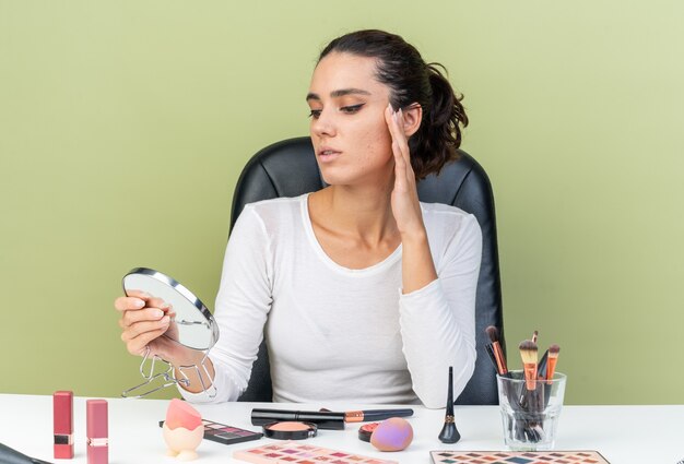 Mulher bonita caucasiana confiante sentada à mesa com ferramentas de maquiagem segurando e olhando no espelho isolado na parede verde oliva com espaço de cópia