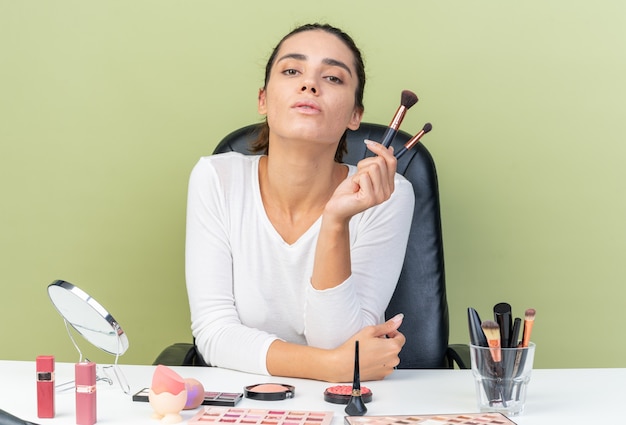 Mulher bonita caucasiana confiante sentada à mesa com ferramentas de maquiagem segurando pincéis de maquiagem