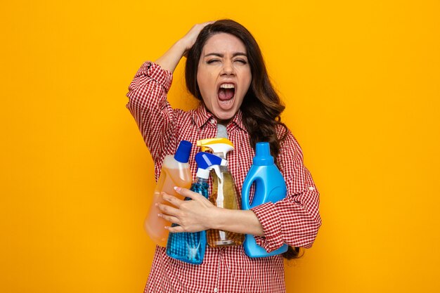 Mulher bonita caucasiana de limpeza irritada segurando sprays e líquidos de limpeza e colocando a mão na cabeça