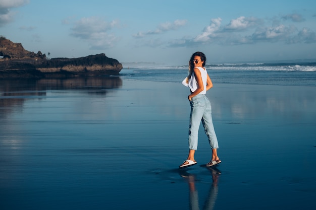Mulher bonita caucasiana em forma de blusa branca e calça jeans refletindo a praia junto ao oceano à luz do sol