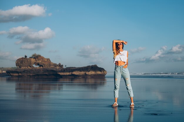 Mulher bonita caucasiana em forma de blusa branca e calça jeans refletindo a praia junto ao oceano à luz do sol