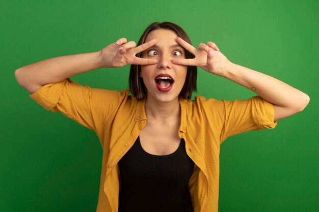 Foto grátis mulher bonita caucasiana engraçada semicerrar os olhos e gesticular sinal de vitória com as duas mãos no verde