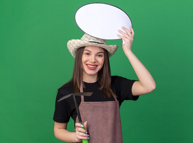 Mulher bonita caucasiana jardineira sorridente com chapéu de jardinagem, segurando o ancinho de enxada e o balão de fala isolado na parede verde com espaço de cópia