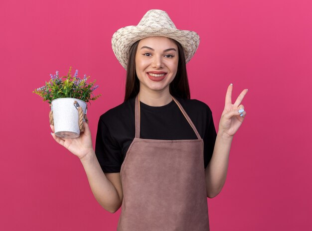 Mulher bonita caucasiana jardineira sorridente com chapéu de jardinagem, segurando um vaso de flores e gesticulando o sinal da vitória, isolada na parede rosa com espaço de cópia