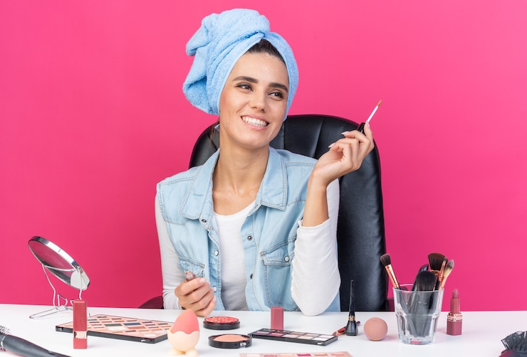 Foto grátis mulher bonita caucasiana sorridente com cabelo enrolado em uma toalha, sentada à mesa com ferramentas de maquiagem segurando brilho labial isolado na parede rosa com espaço de cópia