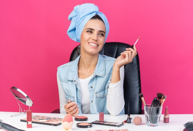 Mulher bonita caucasiana sorridente com cabelo enrolado em uma toalha, sentada à mesa com ferramentas de maquiagem segurando brilho labial isolado na parede rosa com espaço de cópia