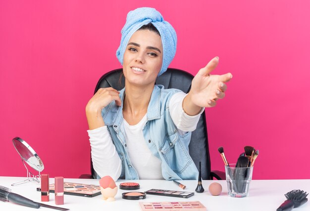 Mulher bonita caucasiana sorridente com o cabelo enrolado em uma toalha, sentada à mesa com ferramentas de maquiagem, estendendo a mão isolada na parede rosa com espaço de cópia