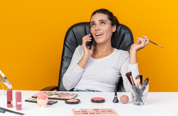 Mulher bonita caucasiana sorridente, sentada à mesa com ferramentas de maquiagem, falando ao telefone, segurando o pincel de maquiagem e olhando para cima