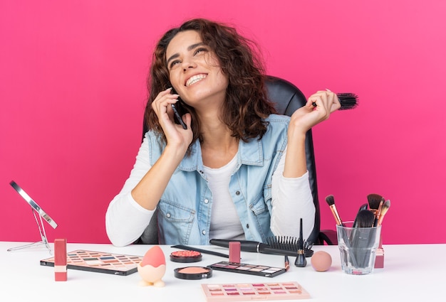 Mulher bonita caucasiana sorridente, sentada à mesa com ferramentas de maquiagem, falando no telefone e segurando o pente isolado na parede rosa com espaço de cópia