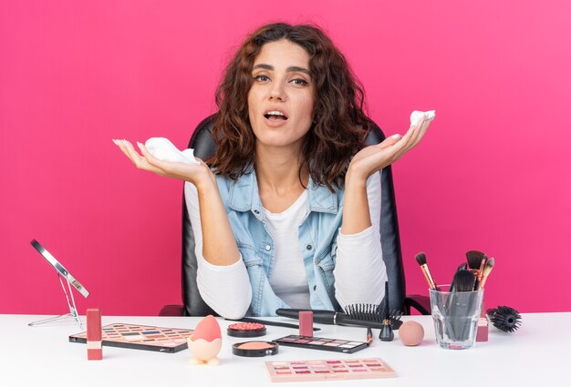 Mulher bonita caucasiana surpresa sentada à mesa com ferramentas de maquiagem segurando mousse de cabelo isolada na parede rosa com espaço de cópia