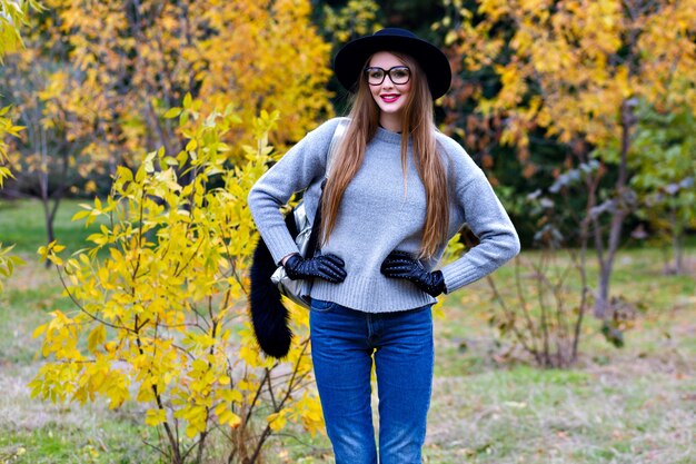 Mulher bonita com cabelo comprido usa jeans e luvas em pé em pose confiante no fundo da natureza. Foto ao ar livre do modelo muito feminino na moda suéter cinza andando no parque em dia de outono.