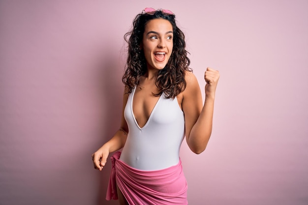 Foto grátis mulher bonita com cabelos cacheados de férias vestindo maiô branco sobre fundo rosa sorrindo com cara de feliz olhando e apontando para o lado com o polegar para cima