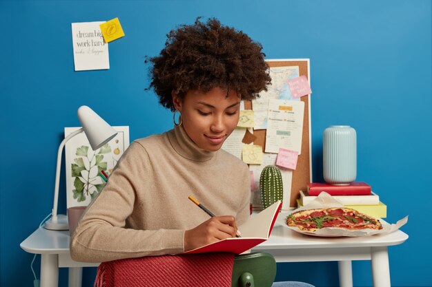 Mulher bonita com corte de cabelo afro faz anotações no bloco de notas, escreve suas próprias ideias, senta-se na cadeira perto da mesa branca com coisas necessárias para o trabalho. Estudo, conceito de educação