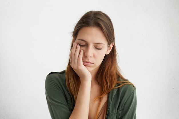 Mulher bonita com expressão sonolenta, parecendo cansada, segurando a mão na bochecha, fechando os olhos de cansaço.