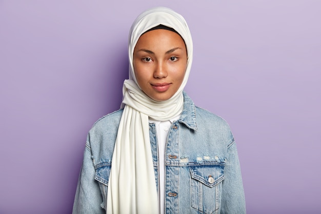 Mulher bonita com pele escura saudável, usa hijab branco, vestida com jaqueta jeans, parece diretamente, isolada sobre a parede roxa. a mulher religiosa tem uma beleza natural. conceito de etnia