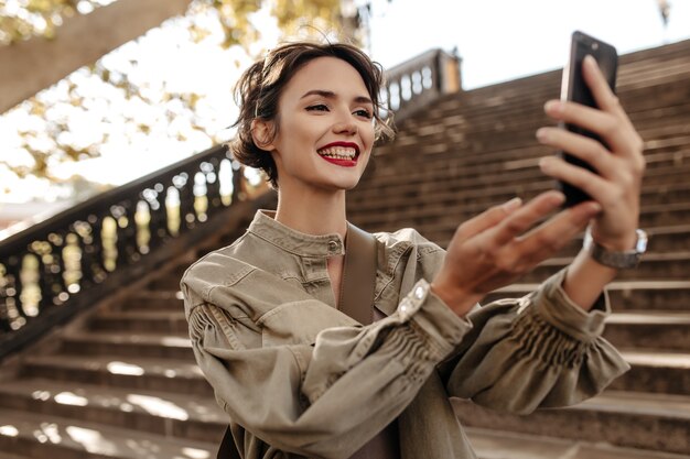 Mulher bonita com penteado curto e lábios vermelhos, sorrindo sinceramente ao ar livre. mulher descolada em jaqueta jeans fazendo selfie