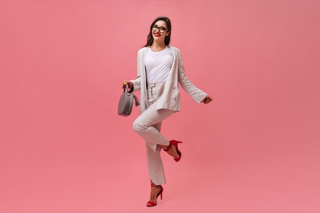 Mulher bonita com roupa bege felizmente posa em fundo rosa. Menina alegre em um terno branco e sapatos vermelhos com bolsa cinza está sorrindo.