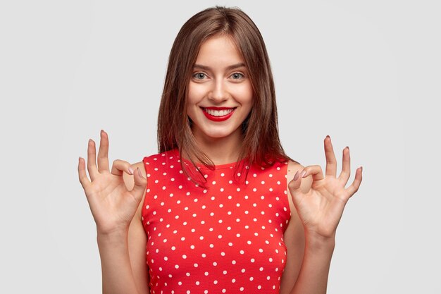 Mulher bonita com sorriso amigável encantador, faz gesto certo, vestida com vestido de bolinhas na moda, mostra aprovação, posa contra uma parede branca. Jovem mulher com lábios vermelhos modelos interiores.