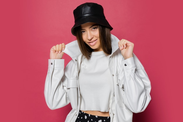 Mulher bonita com um sorriso perfeito, vestida com um boné elegante e uma jaqueta branca, posando em fundo rosa. isolar.