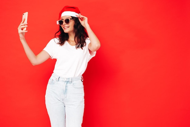 Mulher bonita comemorando o ano novo Feliz mulher linda em roupas casuais tomando selfie na festa de véspera de ano novo Celebração de férias Modelo encantador com chapéu de Papai Noel rindo perto da parede vermelha