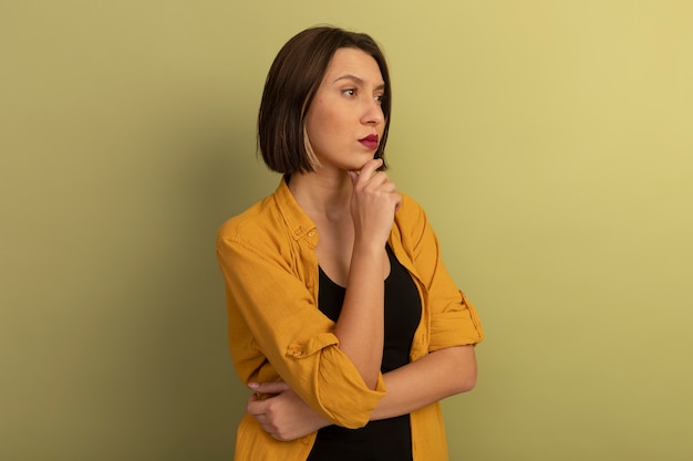 Foto grátis mulher bonita confiante segurando o queixo e olhando para o lado isolado na parede verde oliva