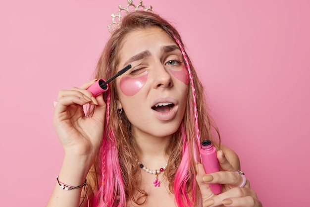 Mulher bonita de cabelos compridos aplica rímel nos cílios faz maquiagem cuida da pele coloca manchas de hidrogel sob os olhos usa coroa e colar se prepara para festa isolada sobre fundo rosa