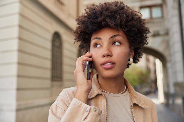 Mulher bonita de cabelos encaracolados faz chamadas de celular passeios ao ar livre na cidade usa jaqueta bege desvia o olhar com expressão pensativa discute as últimas notícias Estilo de vida e conceito de lazer das pessoas