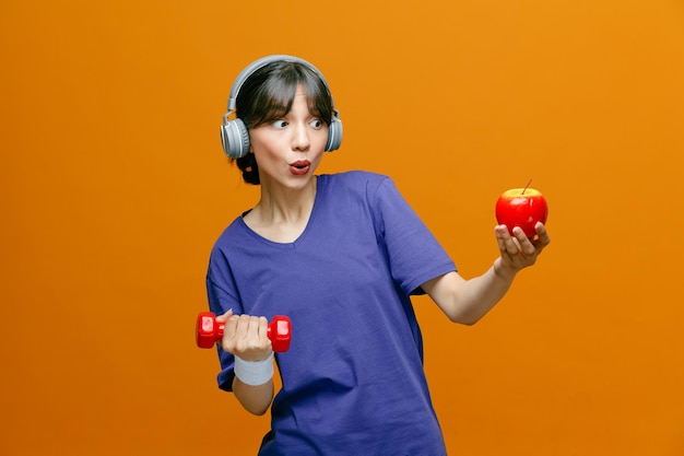 Mulher bonita desportiva em roupas esportivas com fones de ouvido na cabeça fazendo exercícios usando haltere segurando uma maçã olhando para a câmera feliz e positiva em pé sobre fundo laranja