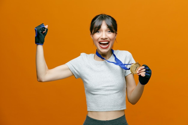 Mulher bonita desportiva em roupas esportivas em luvas com medalha em volta do pescoço, olhando para a câmera feliz e animada, apertando o punho, regozijando-se com seu sucesso em pé sobre fundo laranja