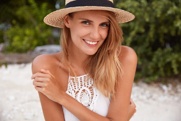 Mulher bonita e alegre com sorriso brilhante e aparência atraente, usa chapéu e vestido de verão, demonstra pele bronzeada perfeita, posa no litoral com expressão positiva. Pessoas e conceito de férias