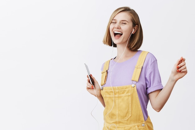 Mulher bonita e alegre dançando com os olhos fechados, despreocupada, segurando um smartphone, ouvindo música nos fones de ouvido