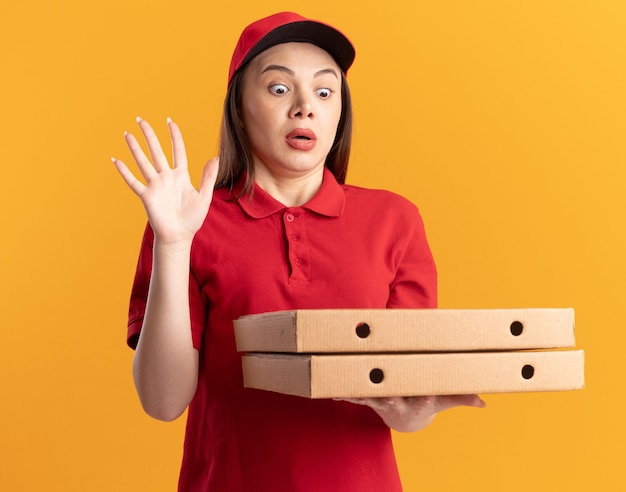 Mulher bonita e ansiosa, entregadora de uniforme, levanta a mão e segura caixas de pizza em laranja