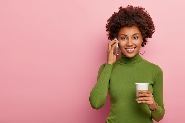 Mulher bonita e atraente com penteado afro, liga para um amigo pelo celular, bebe café para viagem, tem uma conversa agradável, sorri feliz, discute boas notícias, usa um macacão verde casual, posa dentro de casa