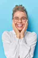 Foto grátis mulher bonita e bonita européia mantém ambas as mãos no rosto sorrisos amplamente vestida com camisa branca óculos transparentes ouve notícias agradáveis sobre um fundo azul parece fofa para a câmera