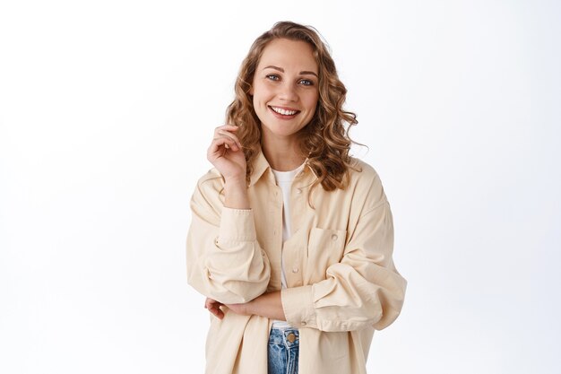 Mulher bonita e cândida com penteado encaracolado, sorrindo e parecendo feliz na frente, em pé otimista contra uma parede branca