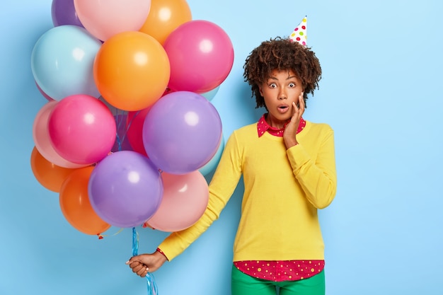 Mulher bonita e chocada segurando balões multicoloridos enquanto posa com um suéter amarelo