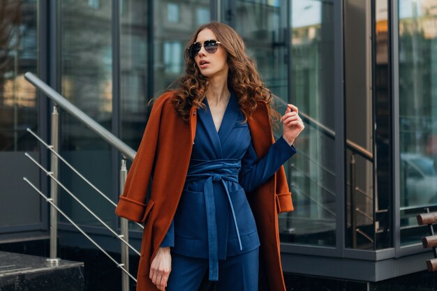 Mulher bonita e elegante andando na rua comercial da cidade urbana, vestida com um casaco marrom quente e terno azul, primavera outono, estilo de moda moderno de rua, usando óculos escuros