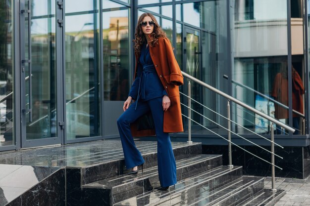 Mulher bonita e elegante andando na rua comercial da cidade urbana, vestida com um casaco marrom quente e terno azul, primavera outono, estilo de moda moderno de rua, usando óculos escuros