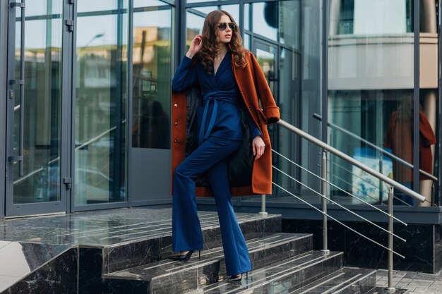 Mulher bonita e elegante andando na rua comercial da cidade urbana, vestida com um casaco marrom quente e terno azul, primavera outono, estilo de moda moderno de rua, usando óculos escuros