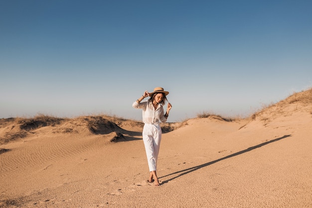 Mulher bonita e elegante caminhando na areia do deserto, com roupa branca e chapéu de palha no pôr do sol