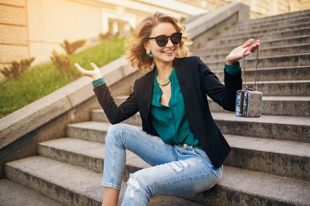 Foto grátis mulher bonita e elegante jovem sentada na escada em uma rua da cidade, vestindo jeans, jaqueta preta, blusa verde, óculos escuros, segurando a bolsa, estilo elegante, tendência da moda de verão, sorrindo