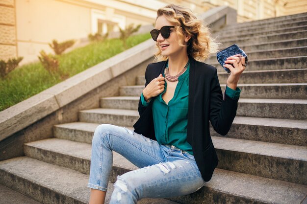 Mulher bonita e elegante jovem sentada na escada em uma rua da cidade, vestindo jeans, jaqueta preta, blusa verde, óculos escuros, segurando a bolsa, estilo elegante, tendência da moda de verão, sorrindo