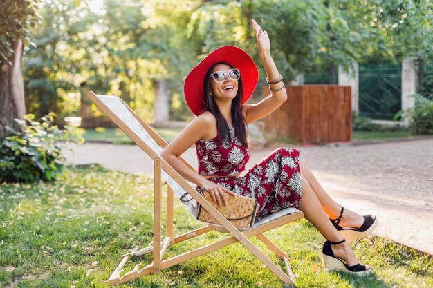 Mulher bonita e elegante sentada na espreguiçadeira com roupa de estilo tropical, acenando com a mão, tendência da moda de verão, bolsa de palha, chapéu vermelho, óculos de sol, acessórios, sorrindo, bom humor, férias