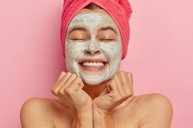 Foto grátis mulher bonita e feliz de olhos fechados, tem máscara de argila no rosto, melhora a aparência, hidrata a pele, sorri amplamente, tem dentes brancos perfeitos, sente-se mimada como em spa, usa toalha no cabelo molhado