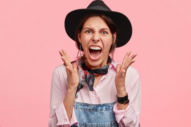 Mulher bonita e irritada, agricultora de chapéu e macacão jeans, gesticula com raiva e mantém a boca bem aberta, tem expressão desesperada, isolada sobre parede rosa