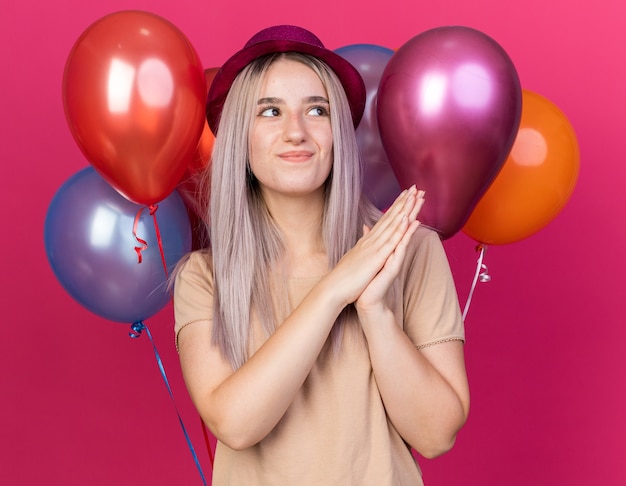 Mulher bonita e jovem impressionada com um chapéu de festa em pé na frente de balões de mãos dadas isoladas na parede rosa