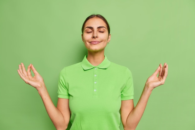 Foto grátis mulher bonita e relaxada que mantém os dedos unidos para meditação faz zen mudra signo respira paciente inala vibrações positivas fecha olhos fechados usa camiseta casual isolada sobre a parede verde