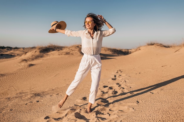 Mulher bonita e sorridente elegante e feliz correndo e pulando na areia do deserto com roupa branca e chapéu de palha no pôr do sol