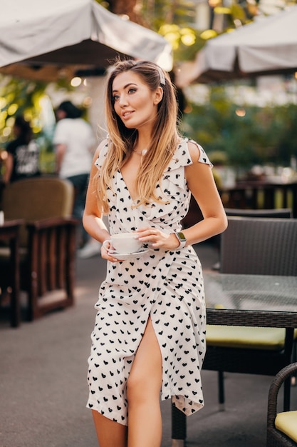 Mulher bonita e sorridente, usando um elegante vestido branco estampado, sentada em um café de rua com uma xícara de cappuccino